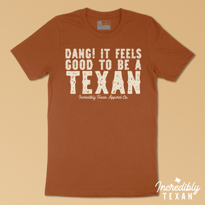 An orange, short sleeve shirt with a light beige print on the front that reads "DANG! IT FEELS GOOD TO BE A TEXAN - Incredibly Texan Apparel Co." in an Old West font.