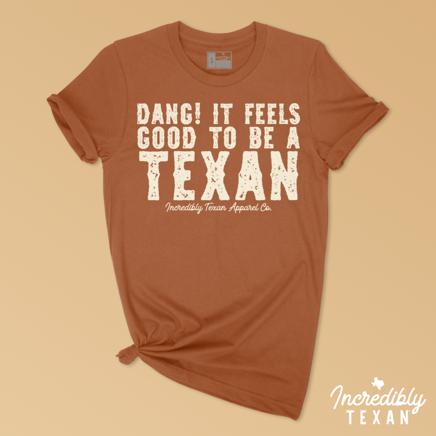 An orange, short sleeve shirt with a light beige print on the front that reads "DANG! IT FEELS GOOD TO BE A TEXAN - Incredibly Texan Apparel Co." in an Old West font.