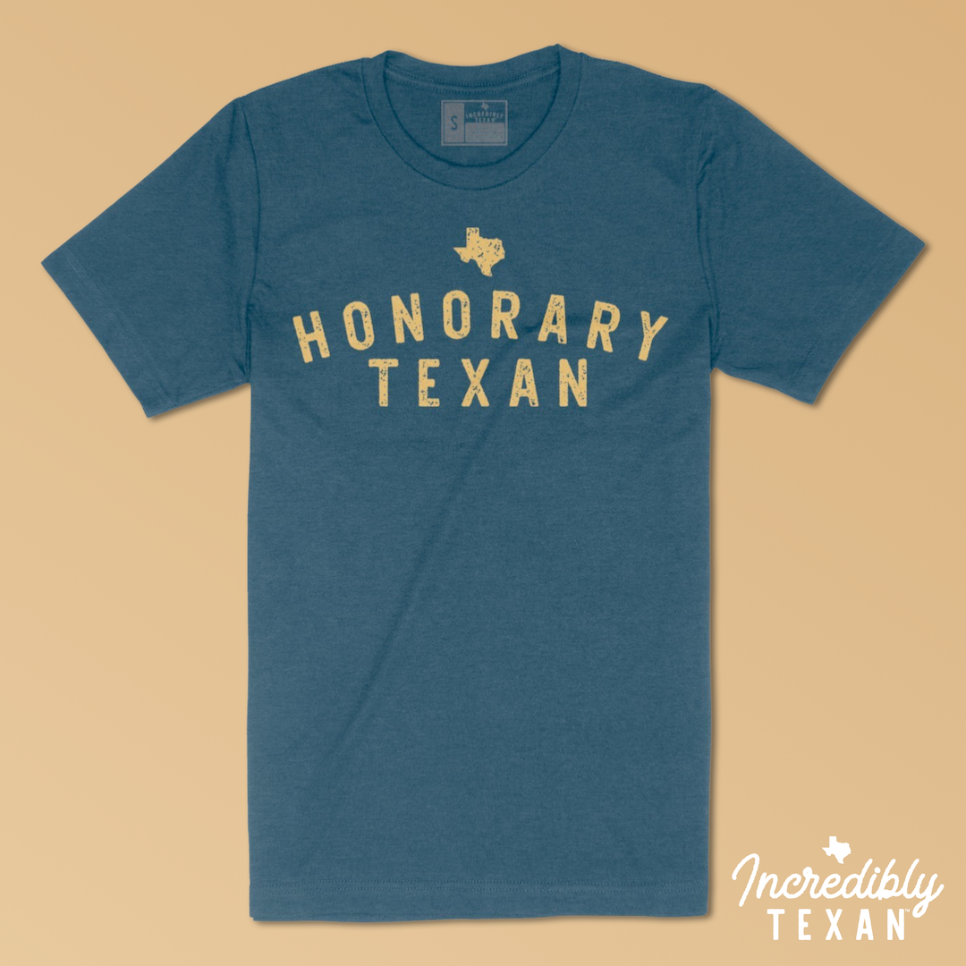 A dark teal blue short sleeve t-shirt with "HONORARY TEXAN" printed in yellow with the state of Texas above.