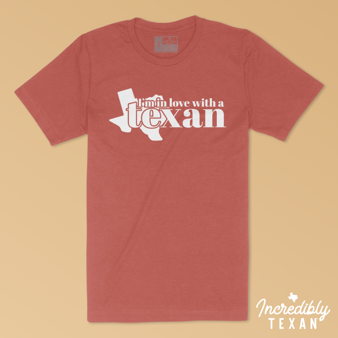 A muted pink short sleeve t-shirt with the words "I'm in Love with a Texan" printed in white with the state of Texas.